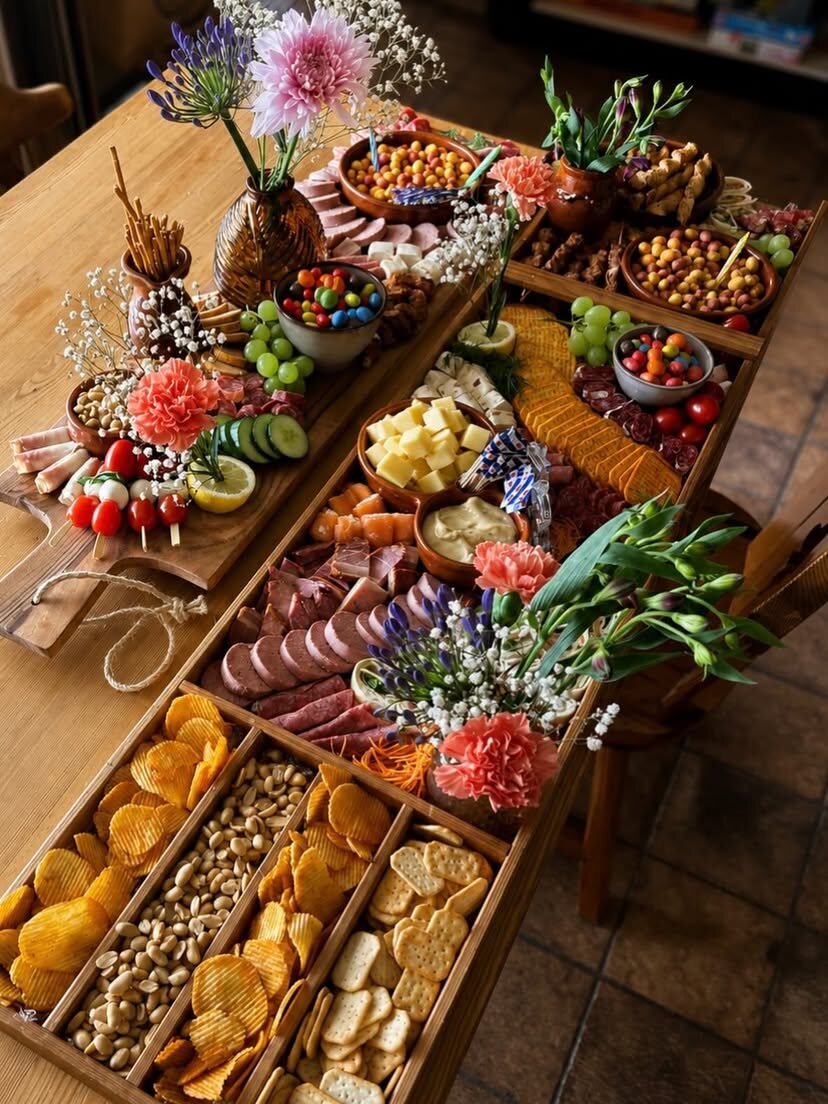 Grazing table van Plankje Geluk over een lange tafel met bloemen, kaas, worst en chips